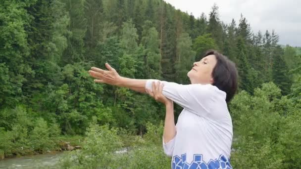 Seniorin mit kurzen dunklen Haaren macht morgens im Freien eine Dehnübung zur Entspannung über Wald- und Gebirgslandschaft — Stockvideo