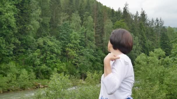 短い黒髪のシニア女性は、森や山の風景の上に午前中にリラクゼーションのためのストレッチ運動を行います — ストック動画