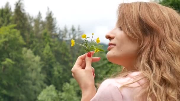 Vacker lockig rödhårig kvinna som innehar en gul vild blomma. Porträtt mot skog och fjäll. Sommarsemester, vårfräschör, mors dag, allergi fri — Stockvideo