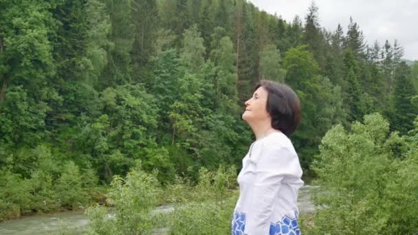 Старшая женщина делает упражнения на растяжку для верхних рук над лесом и горами — стоковое видео