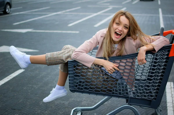 Joven mujer feliz sentada dentro de un carrito de compras y sonriendo en el estacionamiento del supermercado . — Foto de Stock