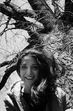 Beyaz ve siyah erken ilkbaharda güneşli bir gün boyunca parkta büyük yaşlı ağacın yanında oturan ceket genç smilling kız outdoos portre kadar