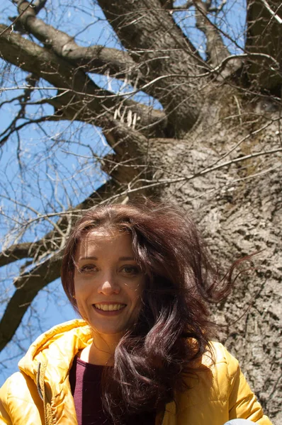 Крупним планом портрет молодої усміхненої дівчини в жовтій куртці, що сидить біля великого старого дерева в парку під час сонячного дня ранньої весни — стокове фото