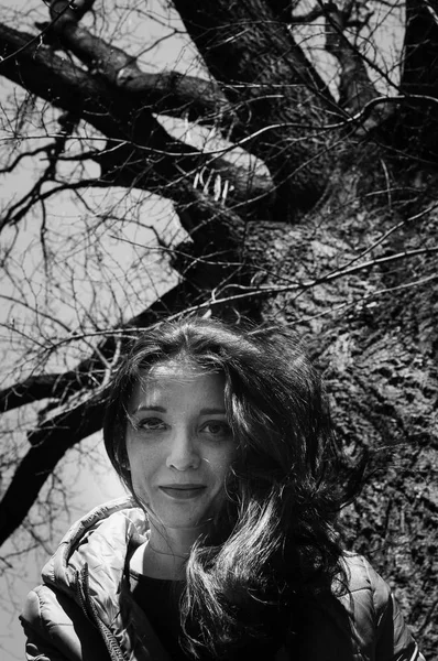 Biały i czarny bliska outdoos portret młodego sfrezowanie dziewczyna w marynarce siedzi w pobliżu dużego starego drzewa w parku w słoneczny dzień wczesną wiosną — Zdjęcie stockowe