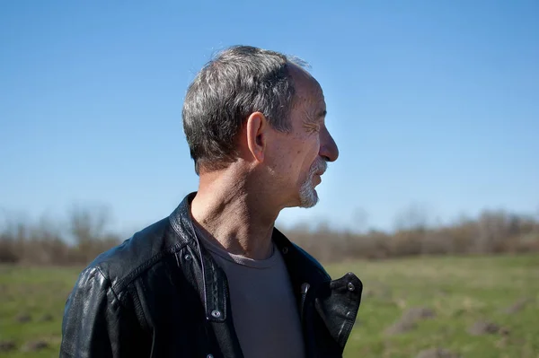 Vista lateral del hombre guapo senior con chaqueta de cuero negro de pie al aire libre en el parque sobre fondo de cielo azul . —  Fotos de Stock