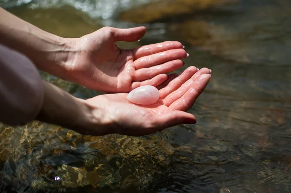 Woman holding in hand a yoni egg. Rose quartz crystal egg on river background. Female health concept
