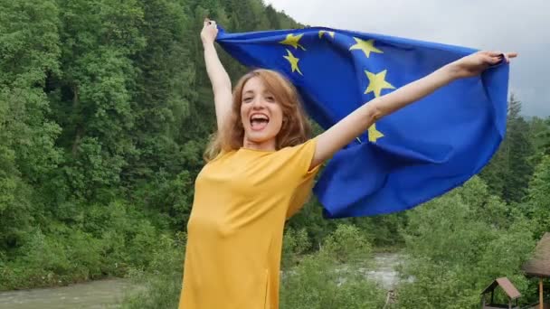 Retrato femenino exterior de una joven patriótica con vestido casual amarillo que sostiene la bandera de la Unión Europea sobre el fondo del bosque verde durante el día de primavera — Vídeos de Stock
