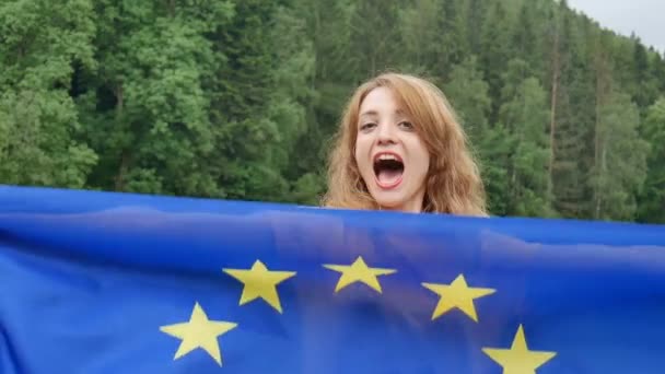 Menina incrível com a bandeira da União Europeia que vai estudar na UE sorrindo e olhando para a câmera no fundo da floresta verde durante o dia de verão ao ar livre . — Vídeo de Stock