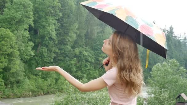 Attractive young woman with umbrella under the spring rain on green forest background. Girl is wearing pink dress and enjoying rainfall, catching a raindrop in her hands. — Stock Video