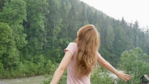 Krásná šťastná dívka stála v letním dešti na pozadí hor pokrytých lesy. Mladá žena šťastně víjí a vychutnává teplý letní déšť