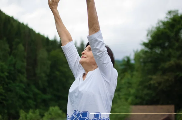 Yaşlı kadın orman ve dağların peyzaj üzerinde dışında üst kollar için bir germe egzersiz yapıyor — Stok fotoğraf
