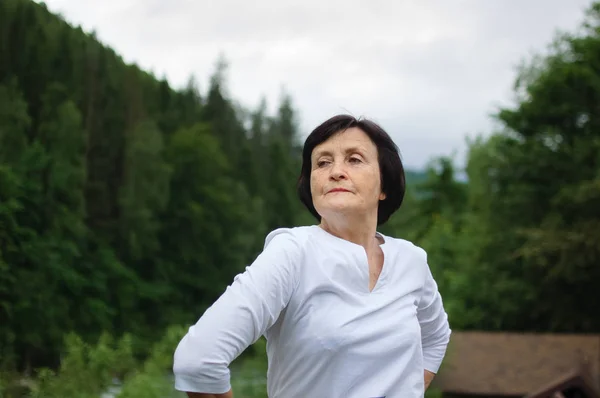 Senior woman doing a stretching exercise for the upper arms outside over landscape of forest and mountains — Stock Photo, Image