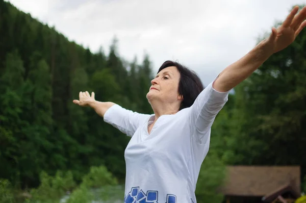Senior woman doing a stretching exercise for the upper arms outside over landscape of forest and mountains — Stock Photo, Image