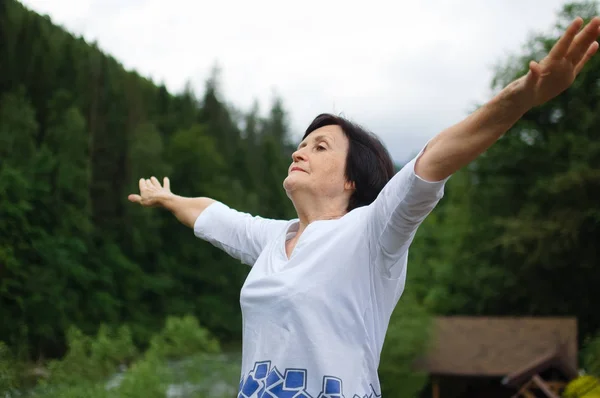 Старшая женщина делает упражнения на растяжку для верхних рук над лесом и горами — стоковое фото