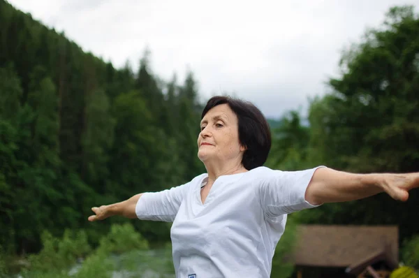 Senior woman doing a stretching exercise for the upper arms outside over landscape of forest and mountains
