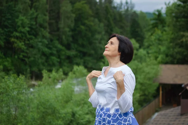 Mujer mayor haciendo un ejercicio de estiramiento para la parte superior de los brazos fuera sobre el paisaje de bosque y montañas —  Fotos de Stock