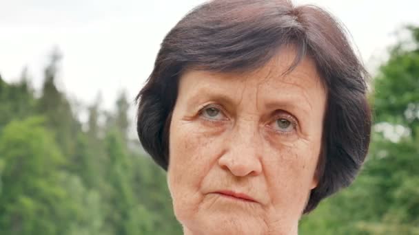 Portrait d'une femme âgée triste et réfléchie aux cheveux bruns courts et aux rides sur son visage sur une colline de montagne avec une forêt verte en arrière-plan — Video