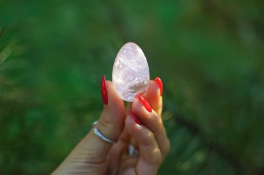 Vumfit, imbuilding veya meditasyon için şeffaf menekşe ametist yoni yumurta tutan kırmızı manikür ile Kadın el. Yeşil arka plan açık havada ellerde Kristal kuvars yumurta