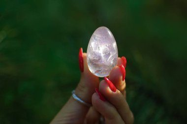 Vumfit, imbuilding veya meditasyon için şeffaf menekşe ametist yoni yumurta tutan kırmızı manikür ile Kadın el. Yeşil arka plan açık havada ellerde Kristal kuvars yumurta