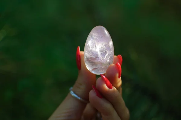 Mano femenina con manicura roja sosteniendo huevo de amatista yoni violeta transparente para vumfit, imbuilding o meditación. Huevo de cuarzo cristal en manos sobre fondo verde al aire libre —  Fotos de Stock