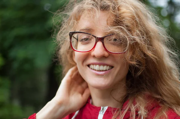 Retrato al aire libre de una joven hermosa mujer con cabello rubio rizado en chaqueta hipster de color rojo oscuro y anteojos mirando a la cámara con sonrisa —  Fotos de Stock