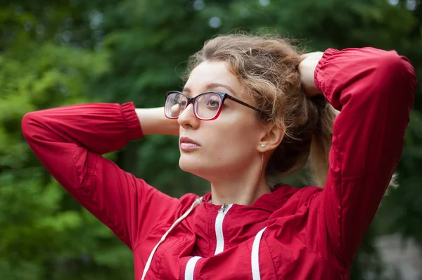 Portrait de fille blonde avec des lunettes hipster en veste rouge foncé faisant queue de cheval à l'extérieur dans le parc urbain — Photo