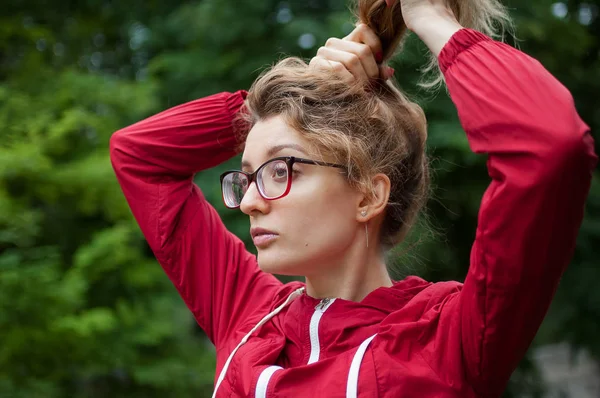도시 공원에서 야외에서 포니 테일을 만드는 어두운 빨간 재킷에 힙스터 안경을 가진 금발 소녀의 초상화 — 스톡 사진
