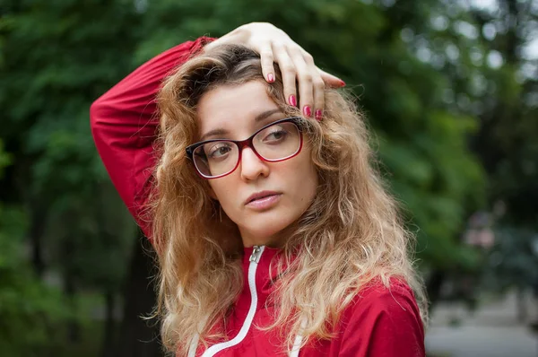 Fiatal szép nő, szőke göndör haja sötét piros csípő kabát és szemüvegek jelentő szabadban a városi parkban reggel — Stock Fotó