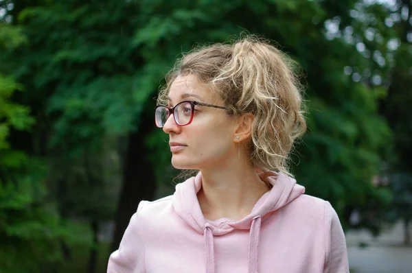 Szabadban női portré fiatal lány szőke haja rózsaszín pulóvert visel csípő szemüvegek a parkban — Stock Fotó