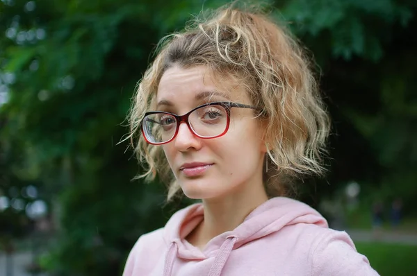 Ao ar livre retrato feminino de menina com cabelo loiro em suéter rosa usando óculos hipster no parque — Fotografia de Stock