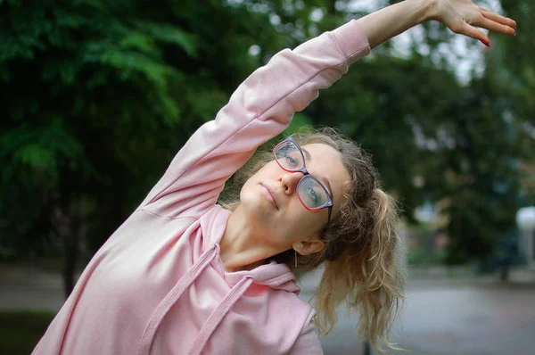 Femme de fitness active en vêtements roses et lunettes s'étire en plein air dans le parc pendant le printemps ou le matin d'été. Concept de mode de vie sain — Photo