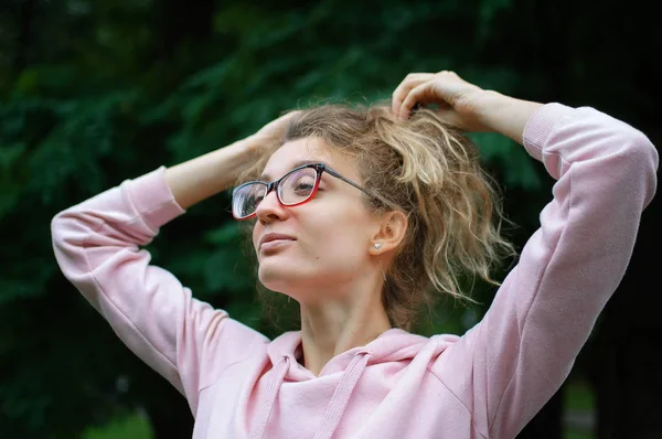 Außenporträt eines blonden Mädchens mit Hipsterbrille in pinkfarbener Kleidung, das draußen im Stadtpark Pferdeschwanz macht — Stockfoto