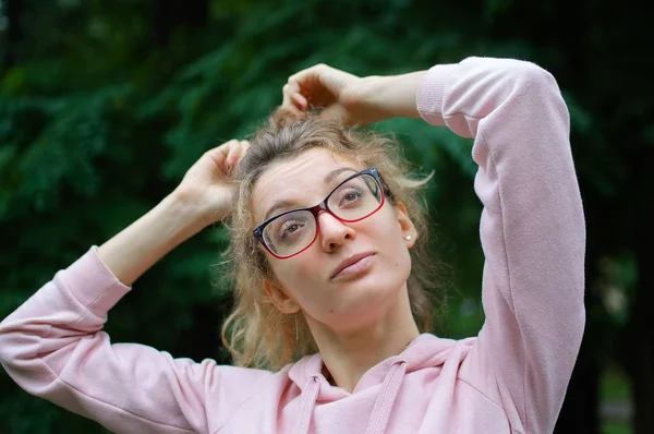 Außenporträt eines blonden Mädchens mit Hipsterbrille in pinkfarbener Kleidung, das draußen im Stadtpark Pferdeschwanz macht — Stockfoto