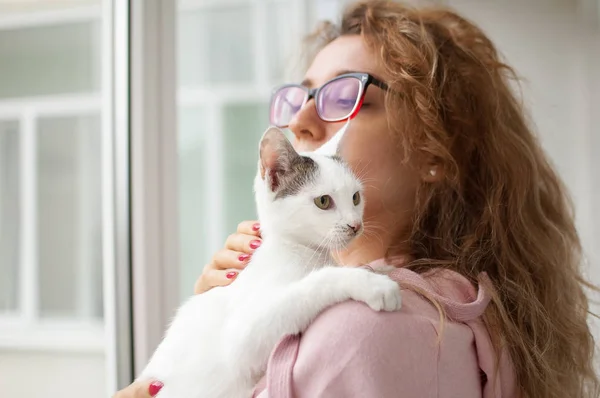 Chica atractiva en ropa rosa y gafas con el gato withe en sus brazos de pie cerca de la ventana en casa. Mascotas concepto propietario — Foto de Stock