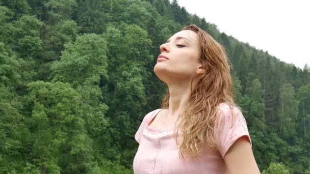 Portrait de femme aux cheveux mouillés blonds et robe rose respirant et se tenant debout sous la pluie froide d'été à l'extérieur dans les montagnes sur fond de forêt verte pendant une journée — Video