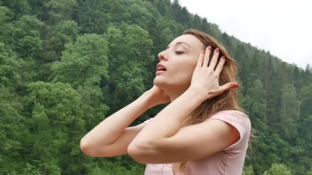 Primer plano de la joven sexy con el pelo mojado rubio y el vestido rosa respirando durante la fría lluvia de verano al aire libre en las montañas sobre el fondo verde del bosque — Vídeo de stock