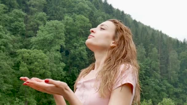 Primer plano de la joven sexy con el pelo mojado rubio y el vestido rosa respirando durante la fría lluvia de verano al aire libre en las montañas sobre el fondo verde del bosque — Vídeos de Stock