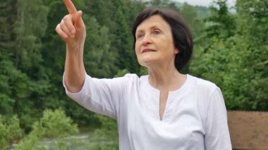 Kısa koyu saçlı ve kırışık yüzü arka planda yeşil orman ile dağ tepeüzerinde dağlara yaptığı gezi sırasında bir şey işaret yaşlı bir kadının Açık hava portresi