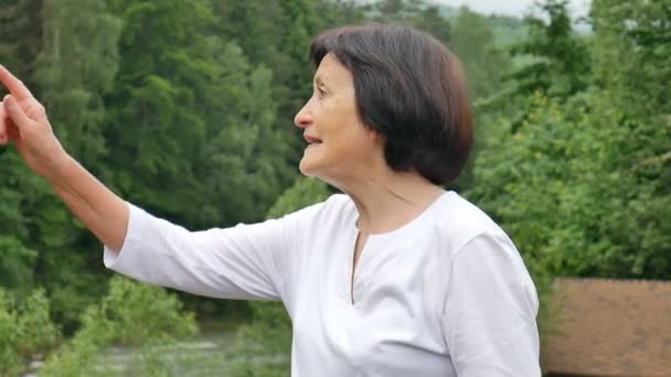 Außenporträt einer älteren Frau mit kurzen dunklen Haaren und faltigem Gesicht, die auf etwas zeigt, während ihrer Reise in die Berge auf einem Hügel mit grünem Wald im Hintergrund — Stockvideo