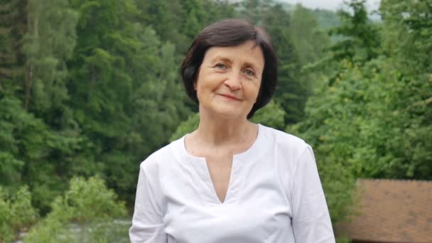 Close up retrato de uma mulher mais velha com cabelo escuro curto e rosto enrugado olhando para a câmera com sorriso na colina da montanha com floresta verde no fundo — Vídeo de Stock
