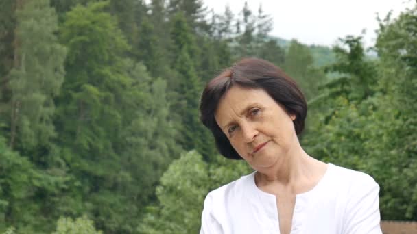 Kısa koyu saçlı ve kırışık yüzü ile hoşnutsuz üzgün yaşlı kadın kadın portresi arka planda yeşil orman ile dağ tepeüzerinde hoşnutsuz açık havada kameraya bakarak — Stok video