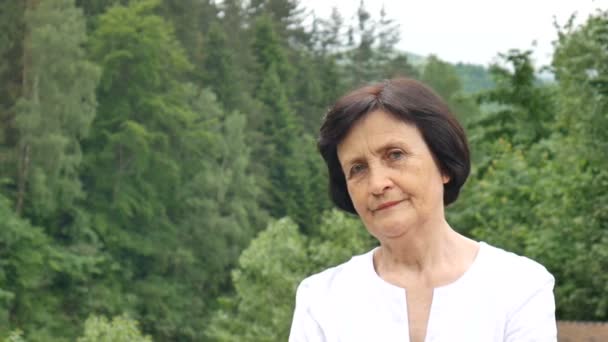 Female portrait of dissatisfied upset older woman with short dark hair and wrinkled face looking at the camera with disapproval outdoors on mountain hill with green forest on background — Stock Video
