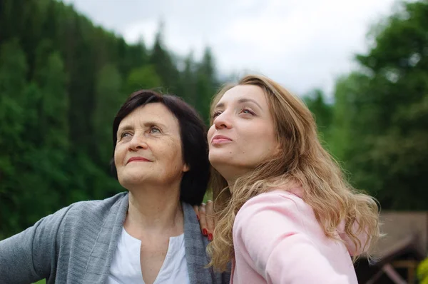 Mother and her adult daughter are travelling and posing together over landscape of forest and mountains. Concept of tenderness, care, family love, relationships.