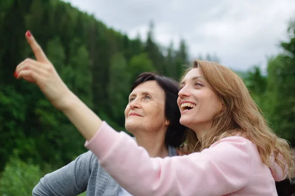 Feliz madre mayor y la hija adulta están viajando y posando juntos sobre el paisaje de bosque y montañas, niña está apuntando a algo. Concepto de viajar en familia —  Fotos de Stock