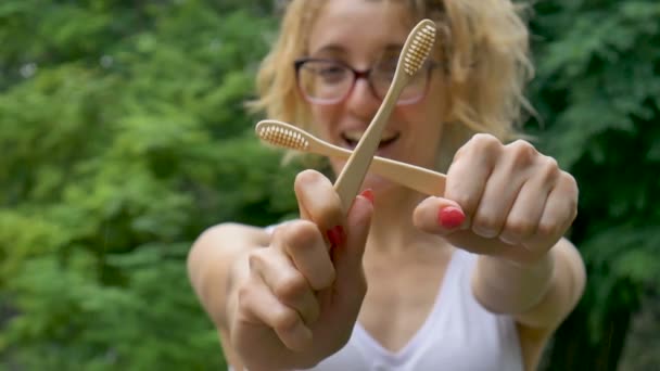 Young beautiful girl with ponytail is holding a useful bamboo toothbrushes outdoors during rainy weather on green tree background. Environmental friendliness and zero waste concept. — Stock Video