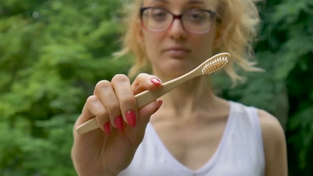 Young beautiful girl with ponytail is holding a useful bamboo toothbrush and looking at the camera outdoors during rainy weather on green tree background. — Stock Video
