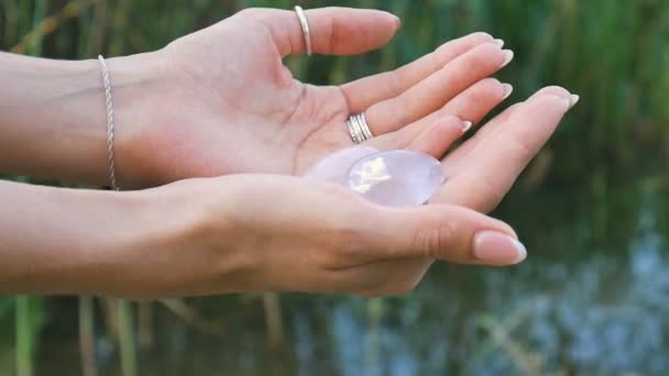 Γυναικείο χέρι κρατώντας ένα τριαντάφυλλο χαλαζία και αμέθυστος κρυστάλλινα αυγά yoni στο φόντο του ποταμού. Υγεία των γυναικών, ενότητα με τις έννοιες της φύσης — Αρχείο Βίντεο