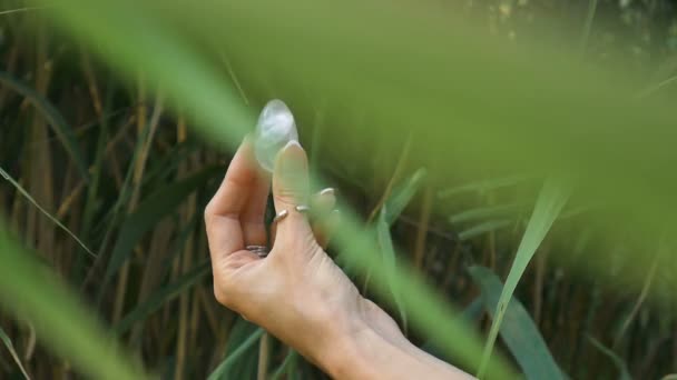Female hand with french manicure holding transparent violet amethyst yoni egg for vumfit, imbuilding or meditation. Crystal quartz egg in hands on green background outdoors. — Stock Video