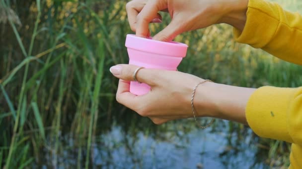 女性の手は、ピンクのシリコーン月経カップで容器を開け、それを示し、湖と緑の自然の背景の中に入れます。女性の健康コンセプト、廃棄物ゼロの選択肢 — ストック動画