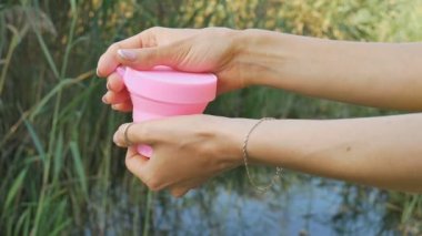 Kadın elleri pembe silikon adet fincan ile bir konteyner açmak, göstermek, ve göl ile yeşil doğal arka plan üzerine koymak. Kadın sağlığı kavramı, sıfır atık alternatifleri